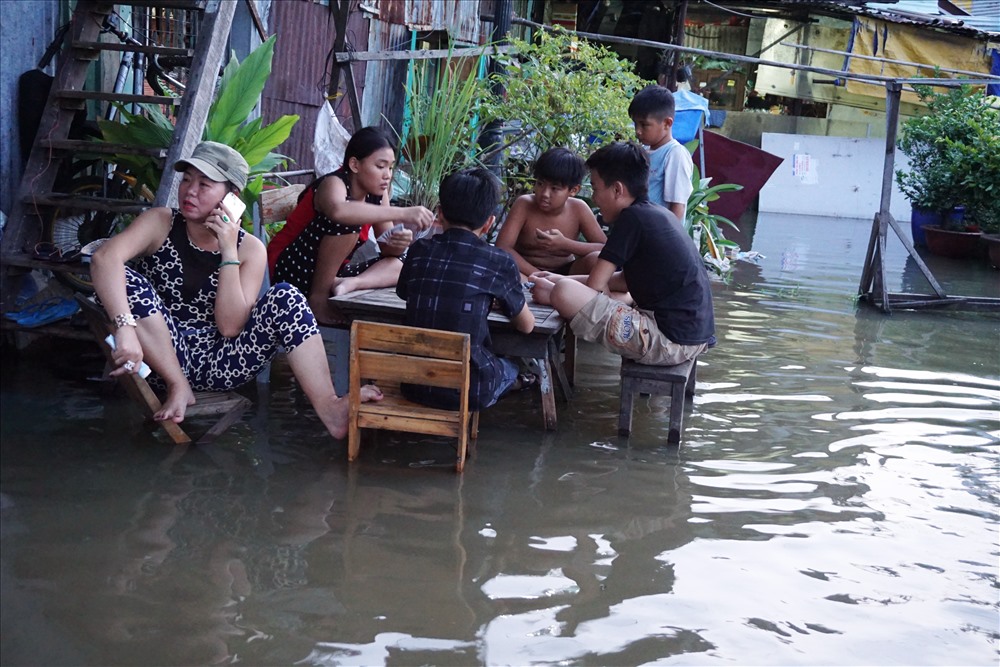 Trong khi người lớn ngồi nhậu, một nhóm trẻ ngồi đáng bài, dưới chân nước ngập lênh láng.