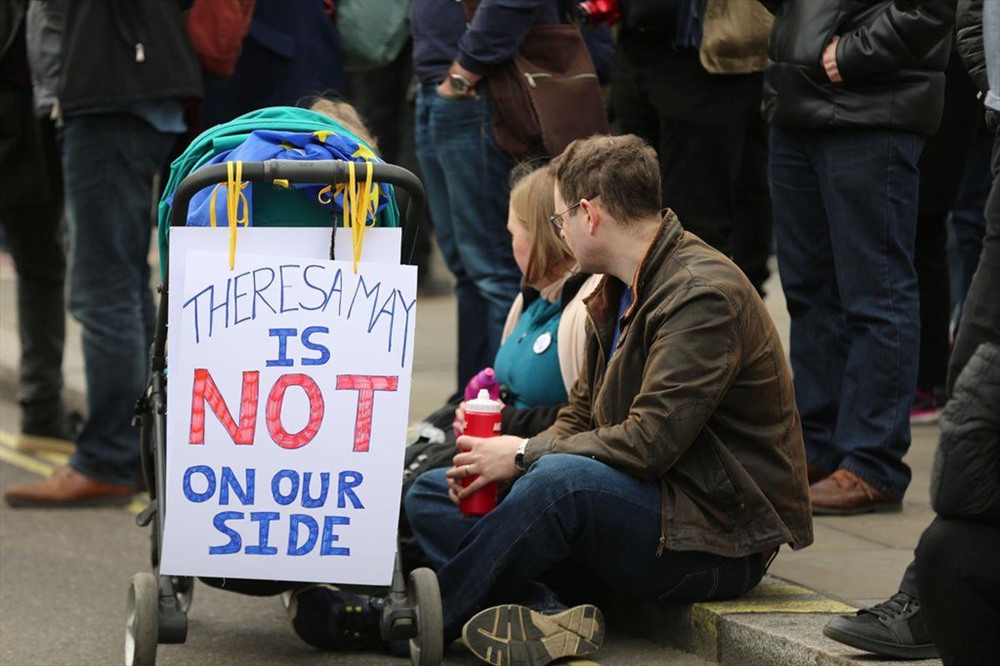 Những người biểu tình ngồi cạnh một chiếc xe đẩy có tấm biển “Theresa May không đứng về phía chúng ta“.